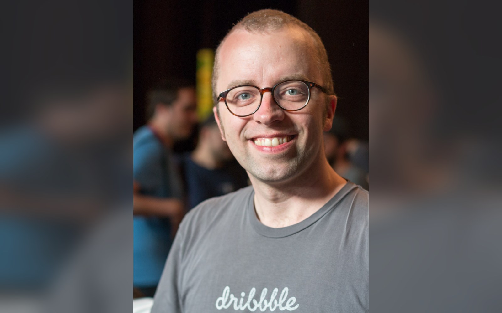 Smiling man with glasses wears a gray dribbble t-shirt at a crowded event